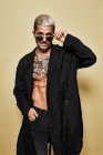 Brutal muscular sexy fit male with tattooed torso wearing black coat and trendy ripped jeans with stylish sunglasses and accessories standing against beige background looking at camera — Stock Photo