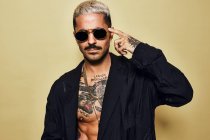 Portrait of brutal muscular sexy fit male with tattooed torso wearing black coat and stylish sunglasses and accessories standing against beige background looking at camera pointing finger to temple — Stock Photo