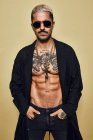 Жорстокий м'язистий сексуальний чоловік з татуйованим торсом у чорному пальто та модними рваними джинсами зі стильними сонцезахисними окулярами та аксесуарами, що стоять на бежевому фоні, дивлячись на камеру — стокове фото