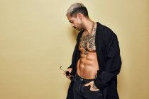 Жестокий мускулистый сексуальный мужчина с татуированным туловищем в черном пальто и модных джинсах со стильными солнцезащитными очками и аксессуарами, стоящими на бежевом фоне, отводя взгляд — стоковое фото