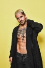 Brutal muscular sexy fit male with tattooed torso wearing black coat and trendy ripped jeans with stylish sunglasses and accessories standing against beige background looking at camera — Stock Photo