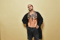 Уверенный в себе стильный мужчина с мускулистым татуированным туловищем в черном пальто и джинсах, смотрящий в камеру на бежевом фоне — стоковое фото