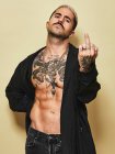Young aggressive provocative male in black coat over naked tattooed torso showing middle finger gesture while standing against beige background — Stock Photo