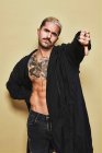 Young provocative male in black coat over naked tattooed torso showing thumbs down gesture in disapproval while standing against beige background — Stock Photo