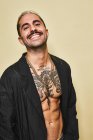 Homme élégant gai confiant avec moustache montrant son torse tatoué musclé portant un manteau noir regardant la caméra sur fond beige — Photo de stock