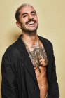 Hombre alegre y seguro con bigote mostrando su musculoso torso tatuado usando un abrigo negro mirando a la cámara sobre un fondo beige - foto de stock