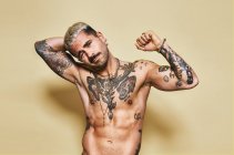 Bonito sexy atraente muscular masculino com várias tatuagens no tronco nu e braços olhando para a câmera enquanto em pé contra o fundo bege — Fotografia de Stock
