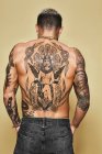Вид ззаду невпізнаваного чоловіка з м'язистим татуйованим тілом у джинсах, що стоять на бежевому фоні — стокове фото
