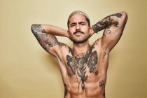 Bonito sexy muscular masculino com várias tatuagens no tronco nu e braços olhando para a câmera enquanto em pé contra o fundo bege — Fotografia de Stock