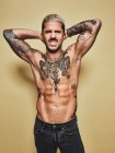 Красивый сексуальный привлекательный мускулистый мужчина с различными татуировками на обнаженном туловище и руках, смотрящий в камеру, корчащий рожи, стоя на бежевом фоне — стоковое фото