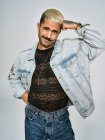 Молодий етнічний чоловік робить похмуре обличчя, дивлячись на камеру в модній джинсовій куртці з квітковим візерунком, стоячи на сірому фоні — стокове фото