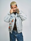Giovane uomo etnico che fa faccia smorfia guardando la fotocamera indossa giacca di jeans alla moda con motivo floreale mentre in piedi su sfondo grigio — Foto stock
