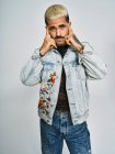 Young ethnic man making grimace doubting face with finger looking at camera wearing trendy denim jacket with floral pattern while standing against gray background — Stock Photo
