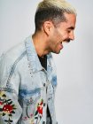 Side view of young unshaven stylish guy in trendy denim jacket laughing while standing against gray background — Stock Photo