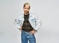 Junger ethnischer Mann mit Fratze und zweifelndem Gesicht, der in trendige Jeansjacke mit Blumenmuster vor grauem Hintergrund in die Kamera blickt — Stockfoto