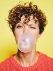 Young curly haired female in orange sweater blowing bubble gum and blinking looking to the camera while standing against yellow background — Stock Photo