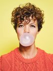 Young curly haired female in orange sweater blowing bubble gum looking to the camera while standing against yellow background — Stock Photo