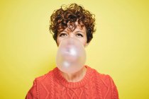 Young curly haired female in orange sweater blowing bubble gum looking to the camera while standing against yellow background — Stock Photo