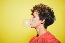 Side view of young pretty curly haired female in orange sweater blowing bubble gum while standing with closed eyes against yellow background — Stock Photo