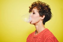 Side view of young pretty curly haired female in orange sweater blowing bubble gum while standing looking away against yellow background — Stock Photo