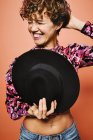 Moda felice modello femminile in possesso di un elegante cappello nero in crop top colorato con stampa floreale in piedi contro sfondo arancione con gli occhi chiusi — Foto stock