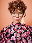 Thoughtful doubtful beautiful curly haired female in trendy eyeglasses and stylish colorful blouse with floral ornament looking at camera against pink background — Stock Photo