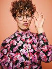 Nachdenklich zweifelhaft schöne lockige Frau in trendiger Brille und stilvoller bunter Bluse mit floralem Ornament blickt in die Kamera vor rosa Hintergrund — Stockfoto