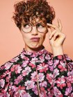 Thoughtful doubtful beautiful curly haired female in trendy eyeglasses and stylish colorful blouse with floral ornament looking away against pink background — Stock Photo