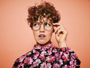 Thoughtful doubtful beautiful curly haired female in trendy eyeglasses and stylish colorful blouse with floral ornament looking away against pink background — Stock Photo