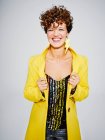 Cheerful woman with sequin top and lightning earring smiling and adjusting stylish yellow coat against gray background — Stock Photo