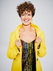 Веселая женщина с блестками сверху и молниеносной серьгой улыбается и регулирует стильное желтое пальто на сером фоне — стоковое фото