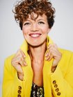 Porträt einer fröhlichen Frau mit Paillettenoberteil und Blitz-Ohrring lächelnd und stylischem gelben Mantel vor grauem Hintergrund — Stockfoto