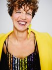 Porträt einer fröhlichen Frau mit Paillettenoberteil und Blitz-Ohrring und stylischem gelben Mantel, die vor grauem Hintergrund in die Kamera lächelt — Stockfoto