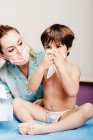 Malato piccolo ragazzo soffiando naso mentre seduto su ospedale letto con femmina infermiera in piedi nelle vicinanze — Foto stock