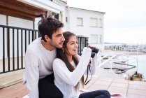 Вид збоку весела романтична пара в повсякденному одязі сидить на терасі з біноклями і насолоджується видом, проводячи разом відпустку на морі — стокове фото