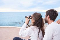Vista lateral do casal romântico alegre em desgaste casual sentado no terraço com binóculos e desfrutando de vista enquanto passam férias juntos à beira-mar — Fotografia de Stock