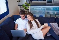 Hochwinkel junger Mann blättert in Laptop und Frau liest interessantes Buch, während sie sich zusammen auf bequemer Couch im modernen Wohnzimmer ausruhen — Stockfoto