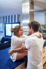 Seitenansicht einer glücklichen jungen Frau, die auf dem Küchentisch sitzt und ihren liebevollen Mann umarmt, während sie den Tag zusammen in einer modernen Wohnung verbringt — Stockfoto