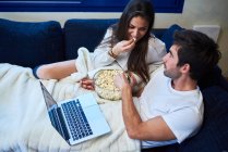 Зверху веселий молодий чоловік і жінка в повсякденному одязі їдять попкорн і дивиться фільм на ноутбук, відпочиваючи разом на затишному дивані вдома — стокове фото