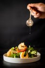 Невпізнаваний кухар додає ложку олії до смачного овочевого салату з креветками на чорному тлі — стокове фото