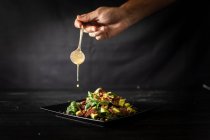 Cuoco irriconoscibile aggiungere cucchiaio di salsa in una porzione gustosa di insalata di avocado appetibile con spinaci freschi e noci serviti su piatto quadrato nero sul tavolo del caffè — Foto stock