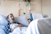 Calm aged man with beard lying under blanket on bed in hospital ward and sleeping — Stock Photo