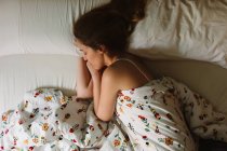 Сверху вид молодой безмятежной женщины с длинными волнистыми волосами в кружевном бюстгальтере, спящей в уютной кровати с белыми простынями и декоративным одеялом — стоковое фото