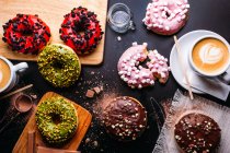 Colocação plana de vários donuts com coberturas doces e barras de chocolate compostas com xícara de cappuccino na mesa preta — Fotografia de Stock