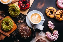 Flat lay of various doughnuts with sweet toppings and chocolate bars composed with cup of cappuccino on black table — Stock Photo