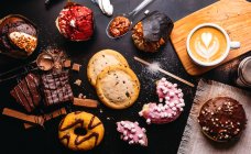 Colocación plana de varias rosquillas con coberturas dulces y barras de chocolate compuestas con taza de capuchino sobre mesa negra - foto de stock