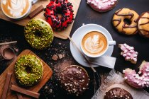 Vários donuts com coberturas doces e barras de chocolate compostas com xícara de cappuccino na mesa preta — Fotografia de Stock
