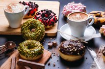 Colocação plana de vários donuts com coberturas doces e barras de chocolate compostas com xícara de cappuccino na mesa preta — Fotografia de Stock