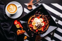 Top view of cappuccino in white mug on table with plate of round waffle with banana and strawberry topped with chocolate sauce and whipped cream — Stock Photo