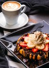 De arriba capuchino en taza blanca sobre mesa con plato de gofre redondo con plátano y fresa rematado con salsa de chocolate y crema batida - foto de stock